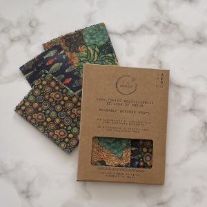 Tres envoltorios de cera de abejas coloridos y estampados se exhiben sobre una superficie de mármol junto a su empaque de cartón marrón etiquetado en español e inglés como Envoltorios de cera de abejas reutilizables.