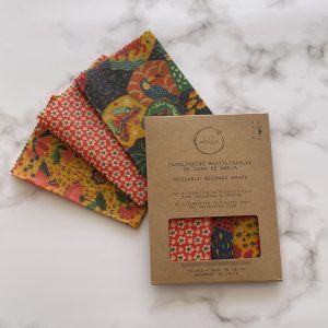 Tres envoltorios de cera de abejas coloridos y estampados se exhiben sobre una superficie de mármol junto a su empaque de cartón marrón etiquetado en español e inglés como Envoltorios de cera de abejas reutilizables.