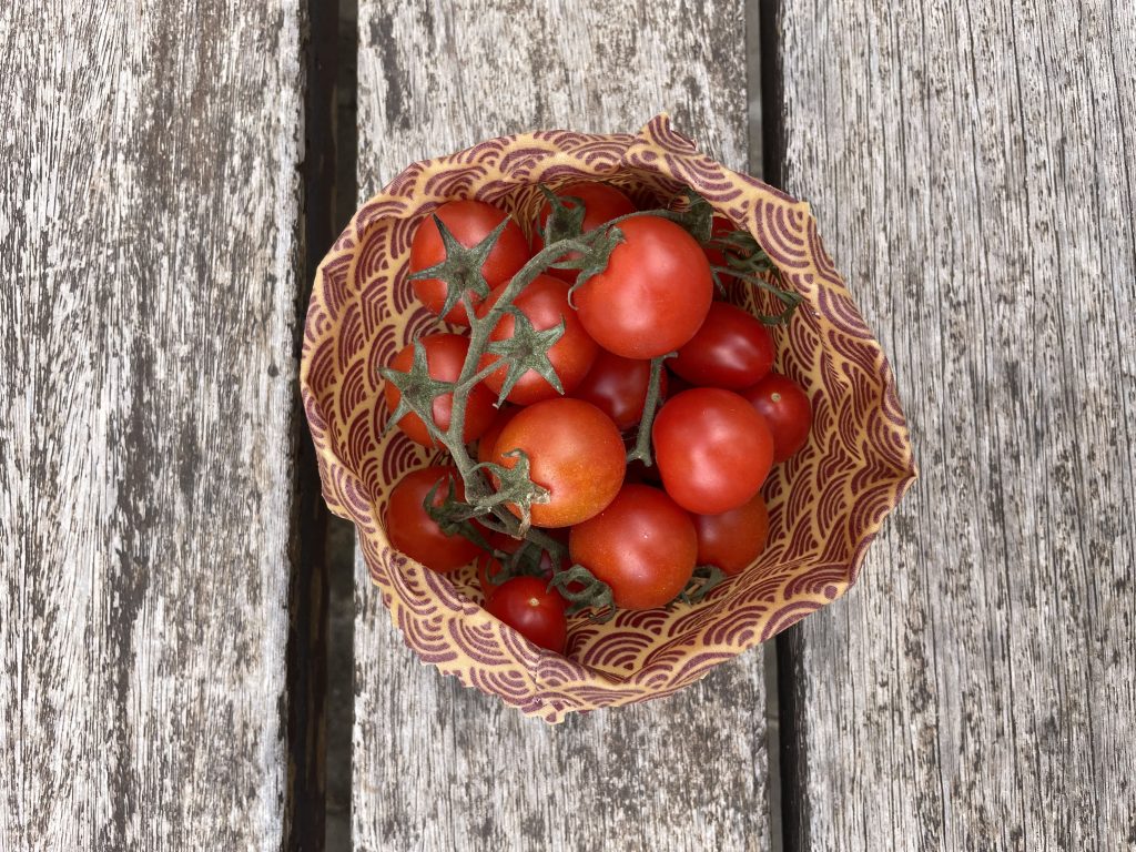 Un cuenco estampado fabricado con una tela encerada para alimentos llena de tomates cherry rojos, algunos todavía en la rama, reposa sobre una superficie de madera desgastada.