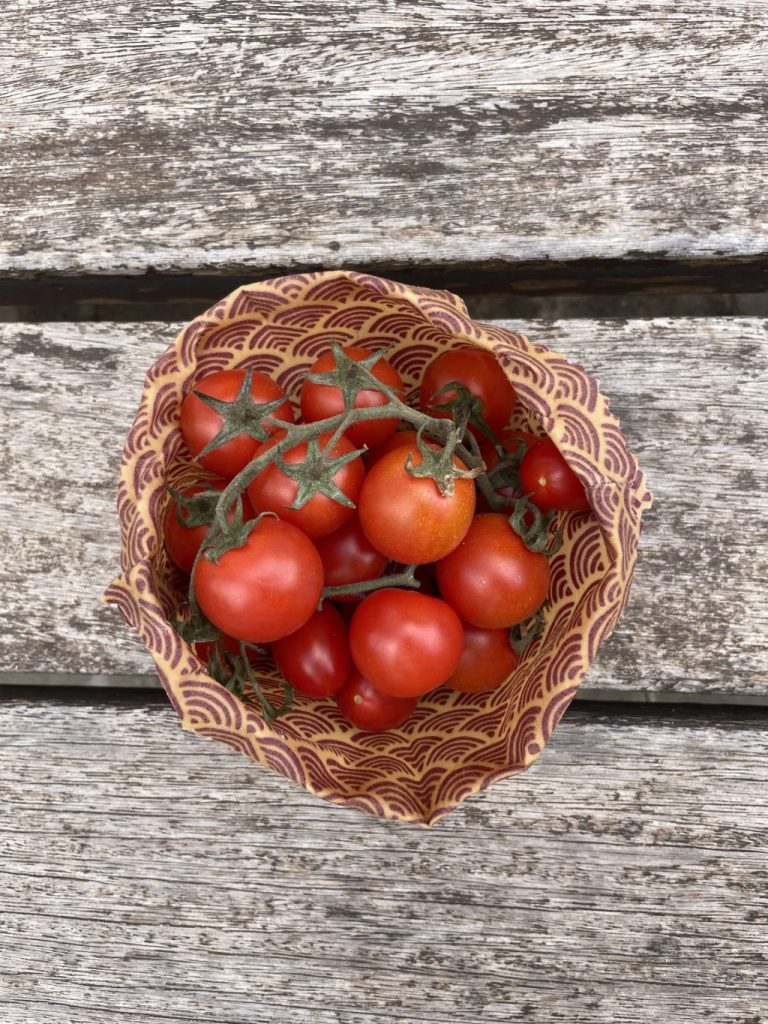 Un cuenco estampado fabricado con una tela encerada para alimentos llena de tomates cherry rojos, algunos todavía en la rama, reposa sobre una superficie de madera desgastada.