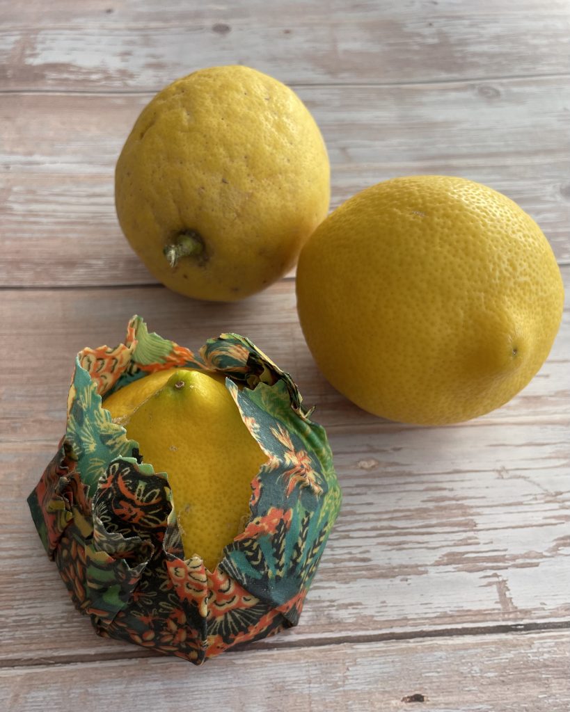 Dos limones enteros se encuentran sobre una superficie de madera clara. En primer plano, un tercer limón está parcialmente envuelto en un envoltorio de cera de abejas con un colorido estampado floral.