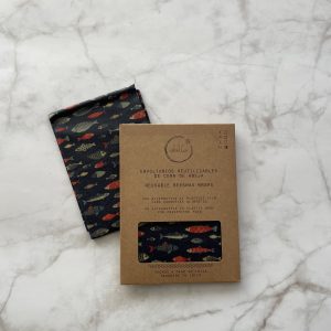 Un envoltorio de cera de abejas colorido y estampado se exhibe sobre una superficie de mármol junto a su empaque de cartón marrón etiquetado en español e inglés como Envoltorios de cera de abejas reutilizables.