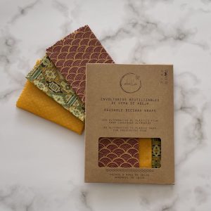 Tres envoltorios de cera de abejas coloridos y estampados se exhiben sobre una superficie de mármol junto a su empaque de cartón marrón etiquetado en español e inglés como Envoltorios de cera de abejas reutilizables.